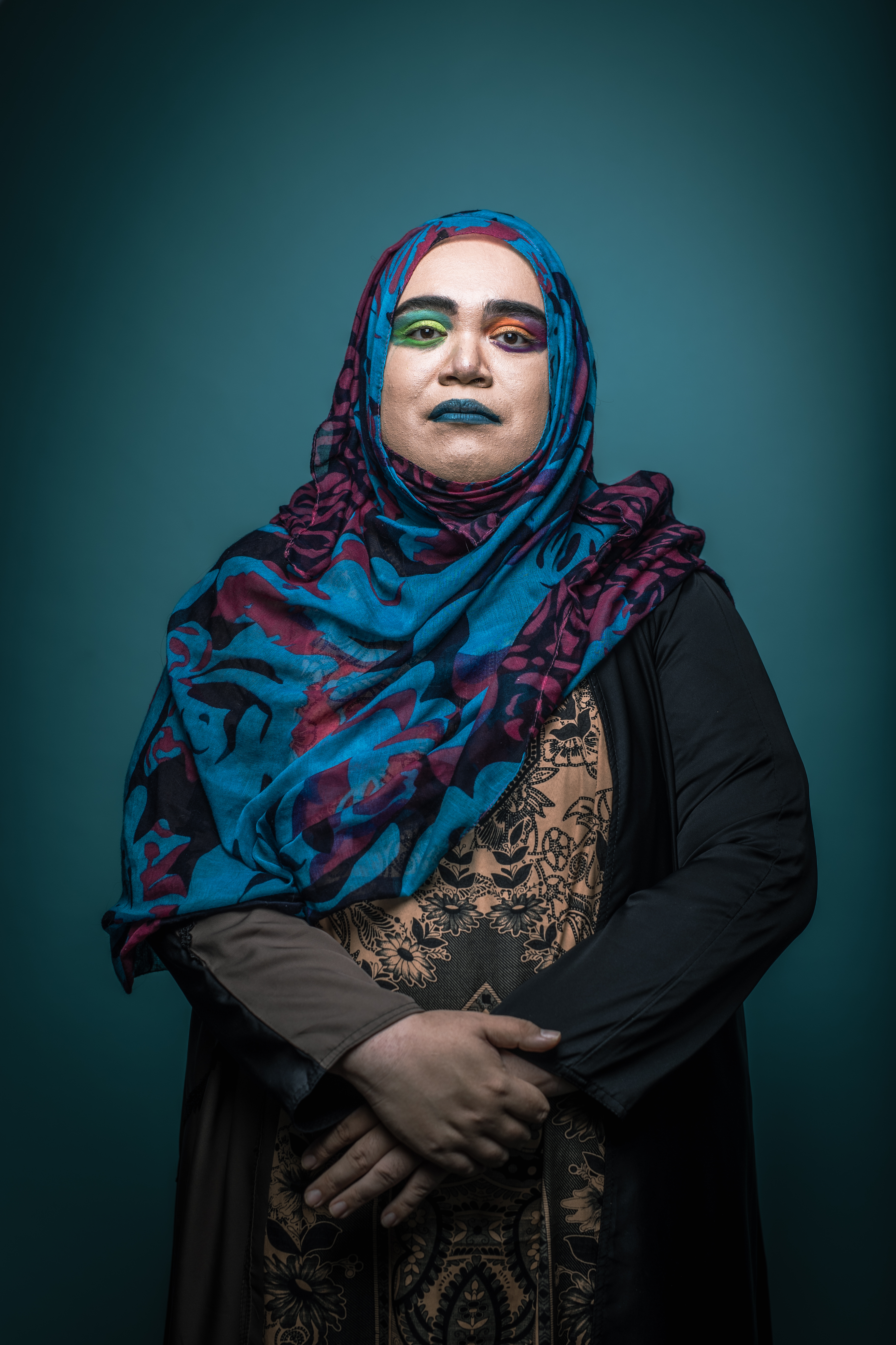Rafiqah Sulaiman Binti Abdullah sitting with her arms in her lap in front of a teal blue background. She is wearing bright coloured rainbow eye shadow and blue lipstick.